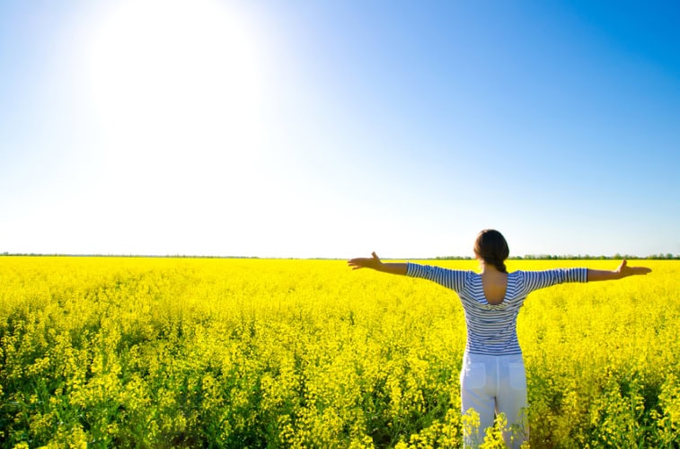 Woman standing in a yellow flower field with arms outstretched, symbolizing mental versatility and openness, under a bright blue sky.