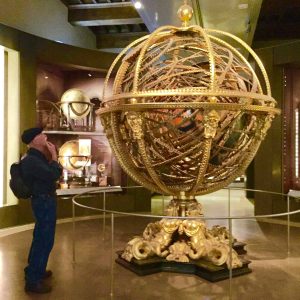 An elaborate, golden globe sculpture with complex interwoven rings and celestial motifs, displayed in a museum setting, symbolizing the interconnectedness of time, knowledge, and the universe.